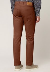 Braune, schmal geschnittene Hose mit Logoschild auf der Rückseite und Fünf-Taschen-Design, kombiniert mit grauen, niedrigen Schuhen, von hinten gezeigt.