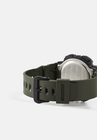 Brassard de montre en silicone vert avec une boucle noire, présentant une surface texturée et des motifs découpés pour la ventilation. Dos de montre en métal visible.
