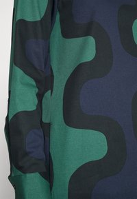 Gros plan sur un tissu avec des formes ondulées abstraites et irrégulières de couleur verte, bleu marine et noire sur une manche de vêtement.