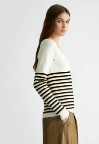 Maglione a righe bianco, nero e oro, con texture a coste, scollatura a V e bottoni decorativi sulla spalla. Maniche lunghe.