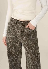 Femme portant un jean large à imprimé léopard et un haut ajusté à manches longues blanc, avec une main dans la poche, coupé au niveau du torse.