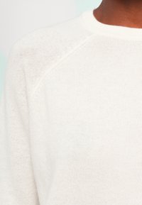 Hvit strikkegenser med rund hals og raglanermer. Myk tekstur, lett materiale og minimalistisk design.
