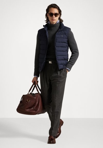 Navy padded vest over a gray turtleneck, gray trousers, and brown leather shoes. Brown leather duffel bag with circular hardware. Dark sunglasses.