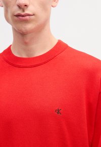 Červený pletený sveter s okrúhlym výstrihom a malým vyšitým logom v čiernej farbe na hrudi. Hladká textúra, neformálny dizajn.