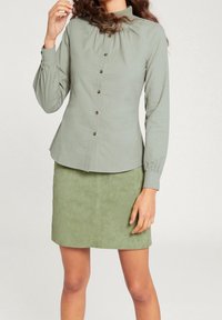 Vaaleanvihreä pitkähihainen nappipaita, jossa on korkea kaulus ja kerrostettu yksityiskohta, yhdistetty samansävyiseen vihreään mokkanahkaseelikseen.