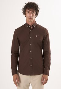 Camicia marrone con bottoni, maniche lunghe e colletto a punta. Presenta un piccolo logo ricamato sul petto e bottoni bianchi.