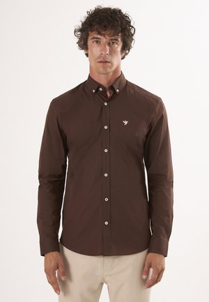 Camisa marrón de botones con mangas largas y cuello puntiagudo. Presenta un pequeño logo bordado en el pecho y botones blancos.