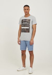 Tricou gri cu text imprimat, șorturi cu model albastru și adidași albi. Design simplu și mâneci scurte, modelul stând în fața unui fundal simplu.