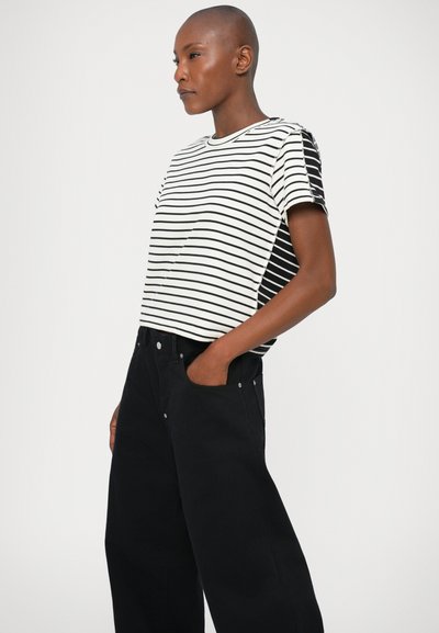 Gestreiftes T-Shirt aus weißer und schwarzer Baumwolle, mit Rundhalsausschnitt und kurzen Ärmeln. Kombiniert mit hochgeschnittenen, schwarzen Schlagjeans.