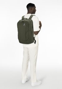 Dunkelgrüner Rucksack aus strapazierfähigem Stoff, mit gepolsterten Tragegurten, mehreren Fächern und einem Logo auf der Vorderseite. Einfaches, kantiges Design.