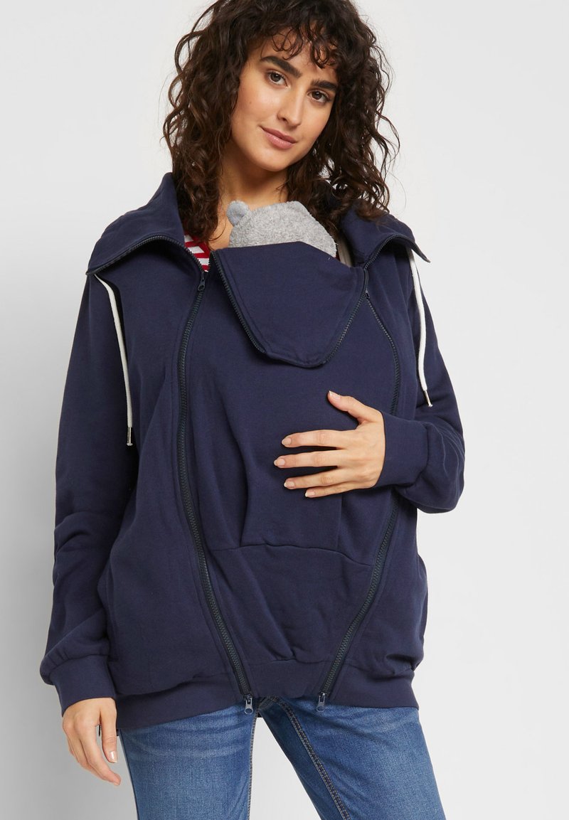 Felpa con zip di colore blu navy realizzata in tessuto morbido. Dotata di una grande tasca frontale con apertura per trasportare un bambino, scollatura regolabile e cordino.