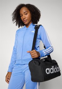 Μπλε ζακέτα φόρμας με φερμουάρ και λευκές ρίγες, σε συνδυασμό με ταιριαστά παντελόνια, κρατώντας μια μαύρη τσάντα duffel με μεγάλο γκρι λογότυπο.