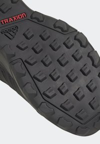 Suola di scarpa atletica nera con motivo di trazione. Presenta una superficie texture e piccoli tasselli rettangolari per la presa. Dettaglio del logo in rosso.