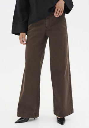 Personne portant un pantalon marron à jambes larges, des talons noirs pointus et un haut noir ample à manches longues sur un fond blanc uni.