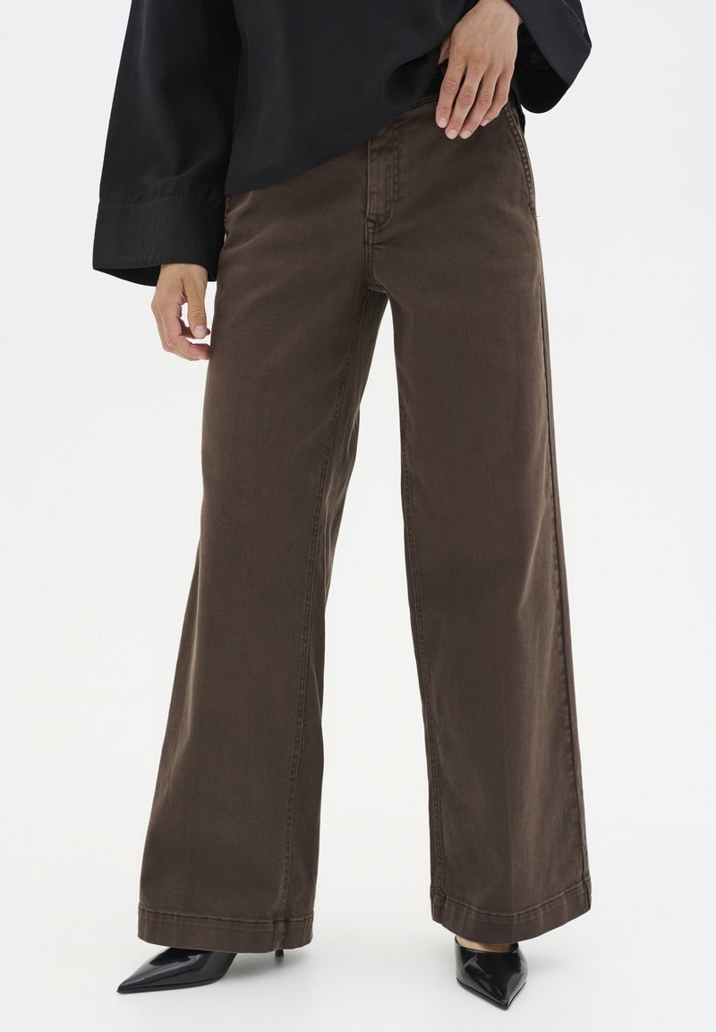 Personne portant un pantalon marron à jambes larges, des talons noirs pointus et un haut noir ample à manches longues sur un fond blanc uni.