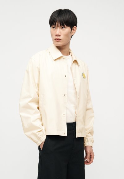 Νεαρός άνδρας φορώντας εκρού τζάκετ με κουμπιά και μικρό μπαλώμα εσπεριδοειδών, άσπρο πουκάμισο από κάτω και μαύρο παντελόνι, στέκεται πλαγιαστά μπροστά σε λευκό φόντο.