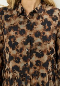 Brązowa i czarna sukienka w kwiatowy wzór z kołnierzykiem zapinanym na guziki, z plisowaną spódnicą i gładką fakturą materiału.