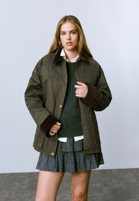 Veste vert olive avec des accents marron, fermeture à boutons et deux poches latérales. Le mannequin porte un pull texturé sombre et une jupe grise superposée.