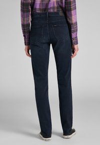 Jeans en denim bleu foncé avec une coupe droite, des poches arrière et une taille mi-haute. Le tissu a une texture lisse, associé à une chemise à carreaux.