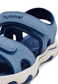 Blå sandal med syntetiskt övermaterial med strukturerade detaljer, kardborreband och en formad vit sula med mönstrad innersula.