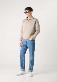 Felpa beige con zip e logo, jeans azzurri e sneakers bianche. Modello indossa occhiali da sole marroni, in piedi contro uno sfondo neutro.