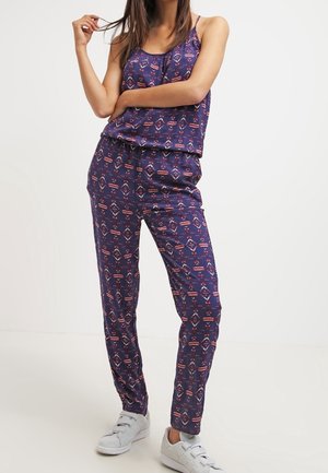 Woman wearing navy blue sleeveless top and matching patterned pants, paired with white sneakers, standing with one arm across her chest.