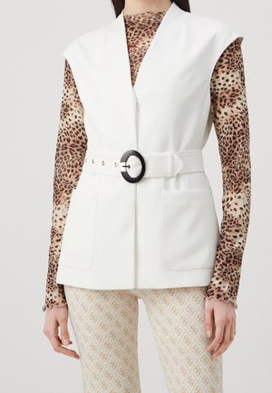 Ärmellose weiße Weste mit Gürtel über einem langärmeligen Oberteil mit Leopardenmuster und gemusterten hellen Hosen an einer stehenden Person.