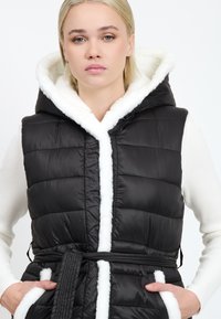 Schwarze Steppweste mit Kapuze und Besatz aus weißem Fleece, mit einem Taillengürtel und gestepptem Design. Taschen sind an den Seiten sichtbar.