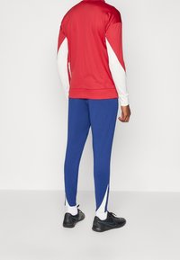 Chándal deportivo con un top rojo que presenta acentos blancos y pantalones azules. El material parece ligero y tiene una textura suave; incluye aberturas laterales.