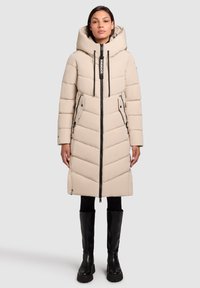 Vrouw in een beige pufferjas tot aan de knie met een grote capuchon, een rits aan de voorkant en zwarte laarzen tot de knie tegen een effen achtergrond.