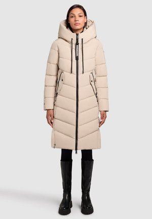 Vrouw in een beige pufferjas tot aan de knie met een grote capuchon, een rits aan de voorkant en zwarte laarzen tot de knie tegen een effen achtergrond.