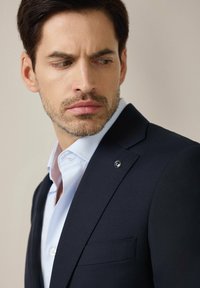 Granatowa marynarka o teksturowanym wykończeniu, z klapami w kształcie notcha i kieszenią piersiową. Nosi się ją na jasnoniebieskiej koszuli z guzikiem. Subtelny detal z logo.