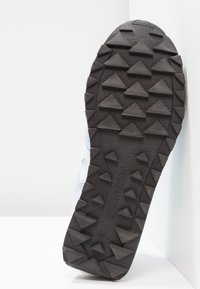 Semelle de chaussure avec des crampons triangulaires en caoutchouc noir disposés en rangées sur un fond blanc, inclinée contre un mur.