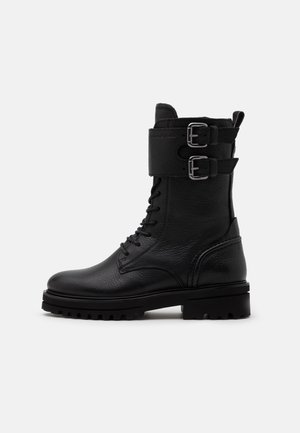 Botte de combat en cuir noir avec fermeture à lacets à l'avant, deux sangles réglables avec boucle en haut, et semelle épaisse crantée.