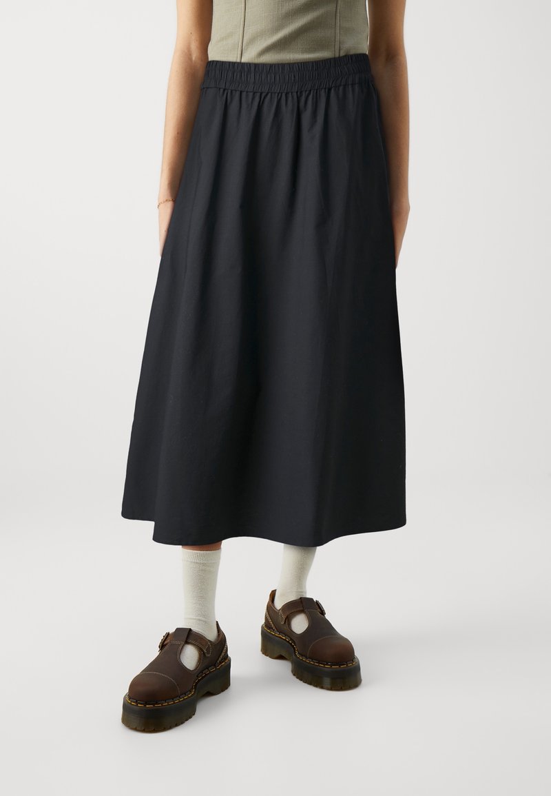 Personne portant une jupe midi noire, un débardeur beige, des chaussettes blanches et des chaussures marron à grosses brides, debout sur un fond uni.