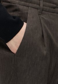 Main insérée dans la poche latérale d'un pantalon marron foncé texturé, portant un vêtement à manches longues noir.