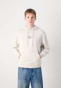 Cremfarbener Hoodie mit einer Fronttasche, Zugbandkapuze und einem kleinen Logo auf der Brust. Weicher Stoff und lockere Passform, kombiniert mit hellblauen Jeans.