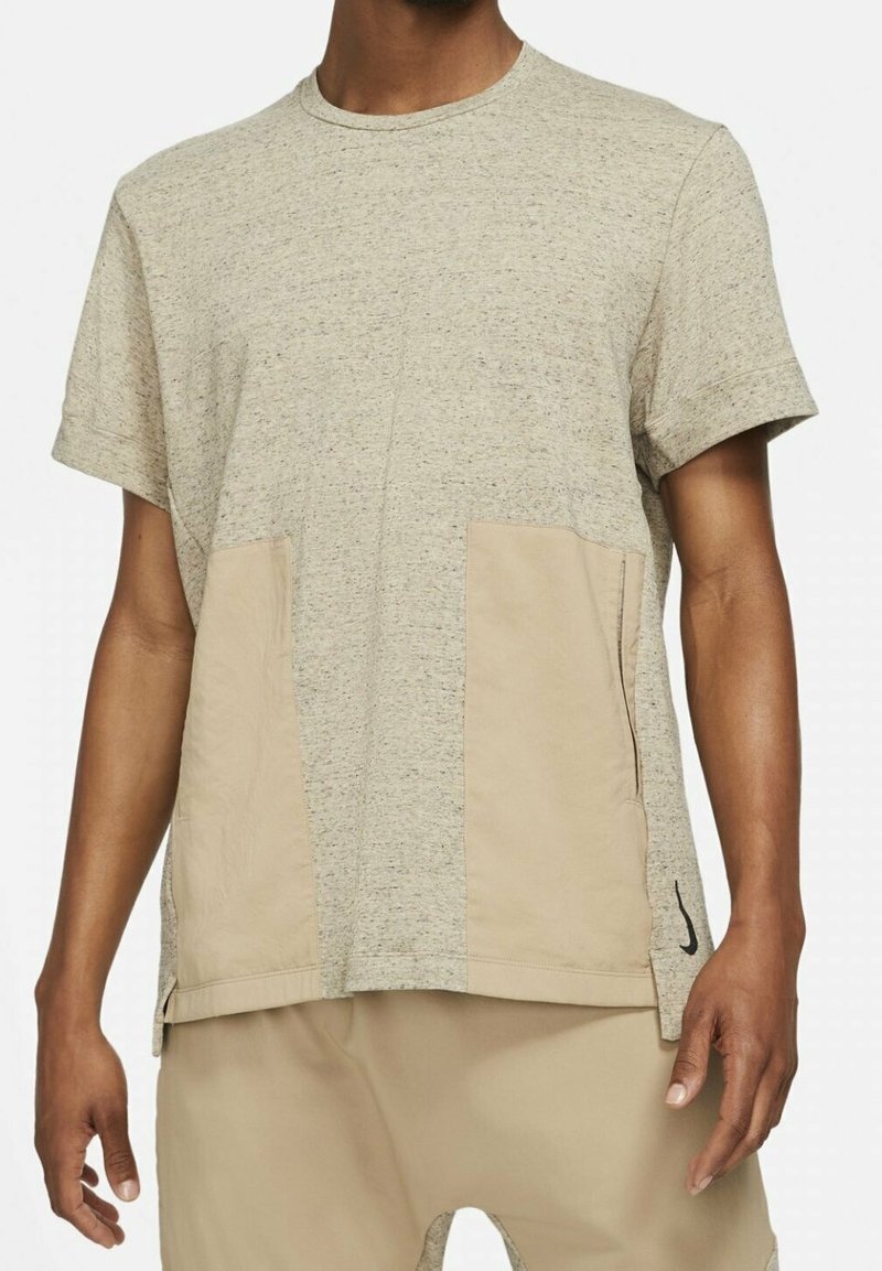 T-shirt gris clair avec une finition texturée. Présente deux panneaux beiges à l'avant et un petit logo noir sur le côté. Manches courtes.