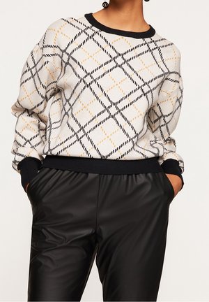 Personne portant un pull blanc avec un motif à carreaux noir et jaune et des poignets noirs, associé à un pantalon en cuir noir, les mains dans les poches.