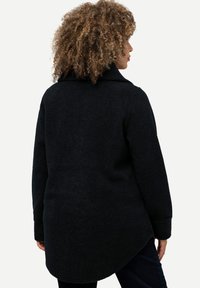 Cappotto nero in misto lana con colletto strutturato, maniche lunghe e orlo arrotondato. Tessuto testurizzato e dettagli delle cuciture laterali visibili.