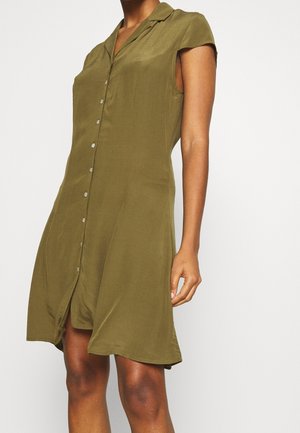 Sleeveless olive green button-up dress with collar, worn by a person against a plain light background.
