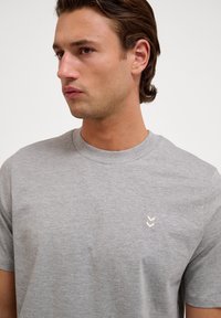Jonge man met kort bruin haar die een lichtgrijs T-shirt met ronde hals en een klein wit Chevron-logo op de borst draagt.