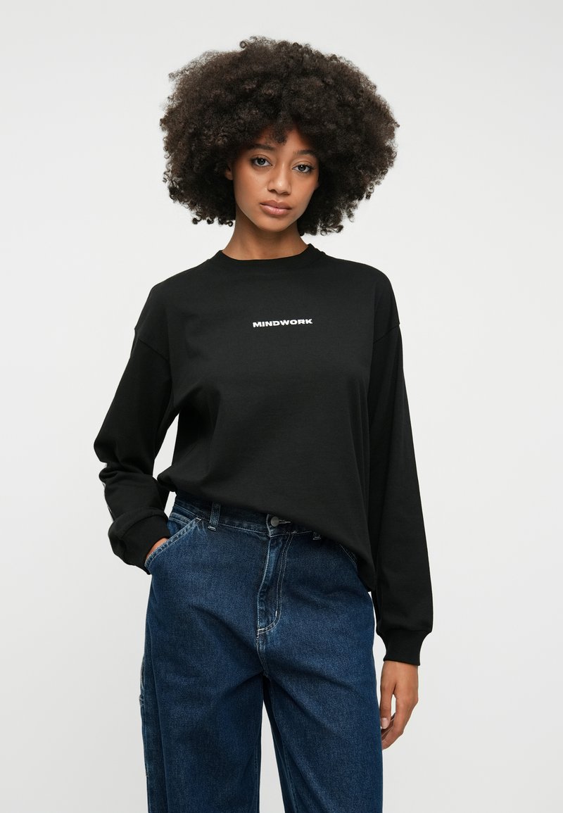 Schwarzes langärmliges T-Shirt mit Rundhalsausschnitt, auf dem "MINDWORK" in weißem Text auf der Vorderseite gedruckt ist. Kombiniert mit blauen Jeans.