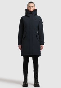 Mørkeblå isoleret parka med høj krave, frontlynlås, trykknaplukninger og to sidelommer. Parret med sorte leggings og snørebåndsstøvler.