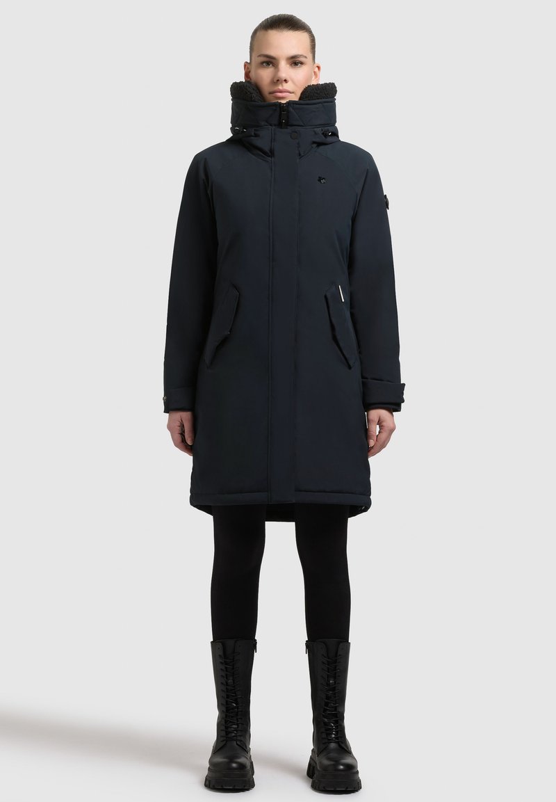 Mørkeblå isoleret parka med høj krave, frontlynlås, trykknaplukninger og to sidelommer. Parret med sorte leggings og snørebåndsstøvler.