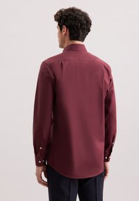 Mann in einem burgunderfarbenen Langarmhemd und dunklen Hosen, von hinten gegen einen einfachen hellen Hintergrund betrachtet.