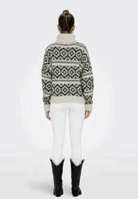 Pull en maille oversize gris clair avec un motif géométrique noir, col montant et poignets côtelés. Associé à un jean blanc et des bottes noires.