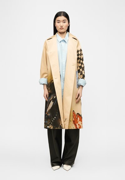 Femme portant un long manteau beige avec des motifs abstraits noirs, bruns et à carreaux, une chemise à rayures bleues claires, un pantalon noir et des chaussures blanches à bout pointu.