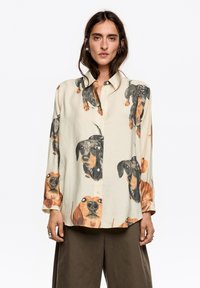 Koszula na guziki z kołnierzykiem w kremowym kolorze, zdobiona wieloma ilustracjami jamników w różnych pozach i kolorach. Luźny krój z długimi rękawami.