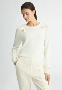 Pull en tricot blanc avec des manches côtelées et des bretelles décorées de boutons. Associé à un pantalon tailleur de couleur claire.
